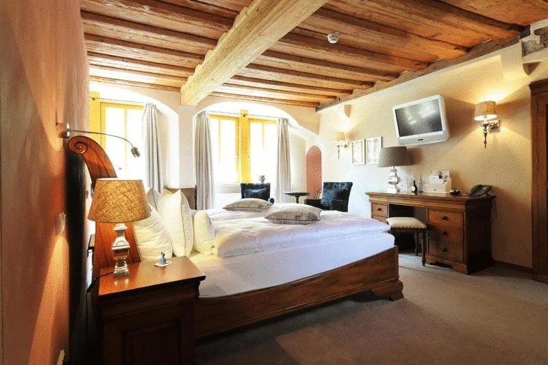 A cozy hotel room with a wooden ceiling, a large bed with white linens, a bedside table with a lamp, two windows with white curtains, and a sitting area with two armchairs. There's a wall-mounted TV, a wooden desk, and a small wooden wardrobe.