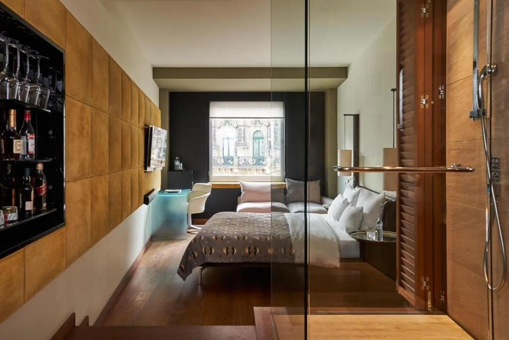 A modern hotel room with a glass wall separating the sleeping area from the bathroom shower.