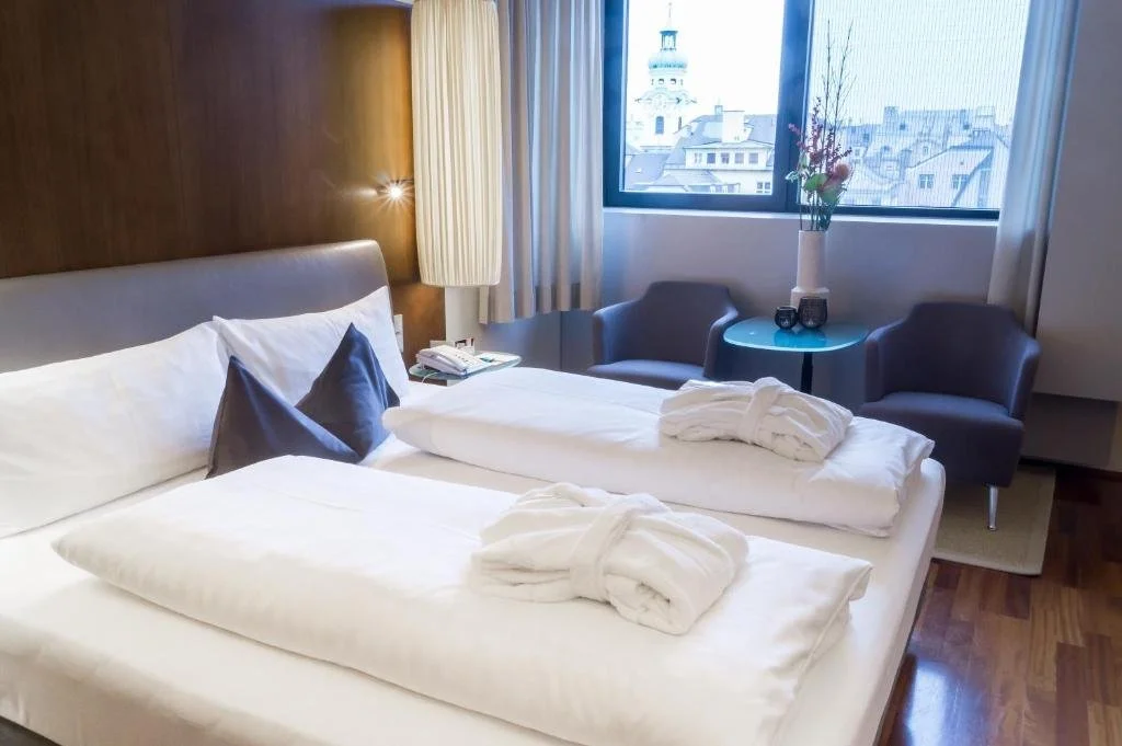 A hotel room with a large bed, two gray chairs near a window with city view, a small round table with a vase of flowers, and folded bathrobes on the bed.