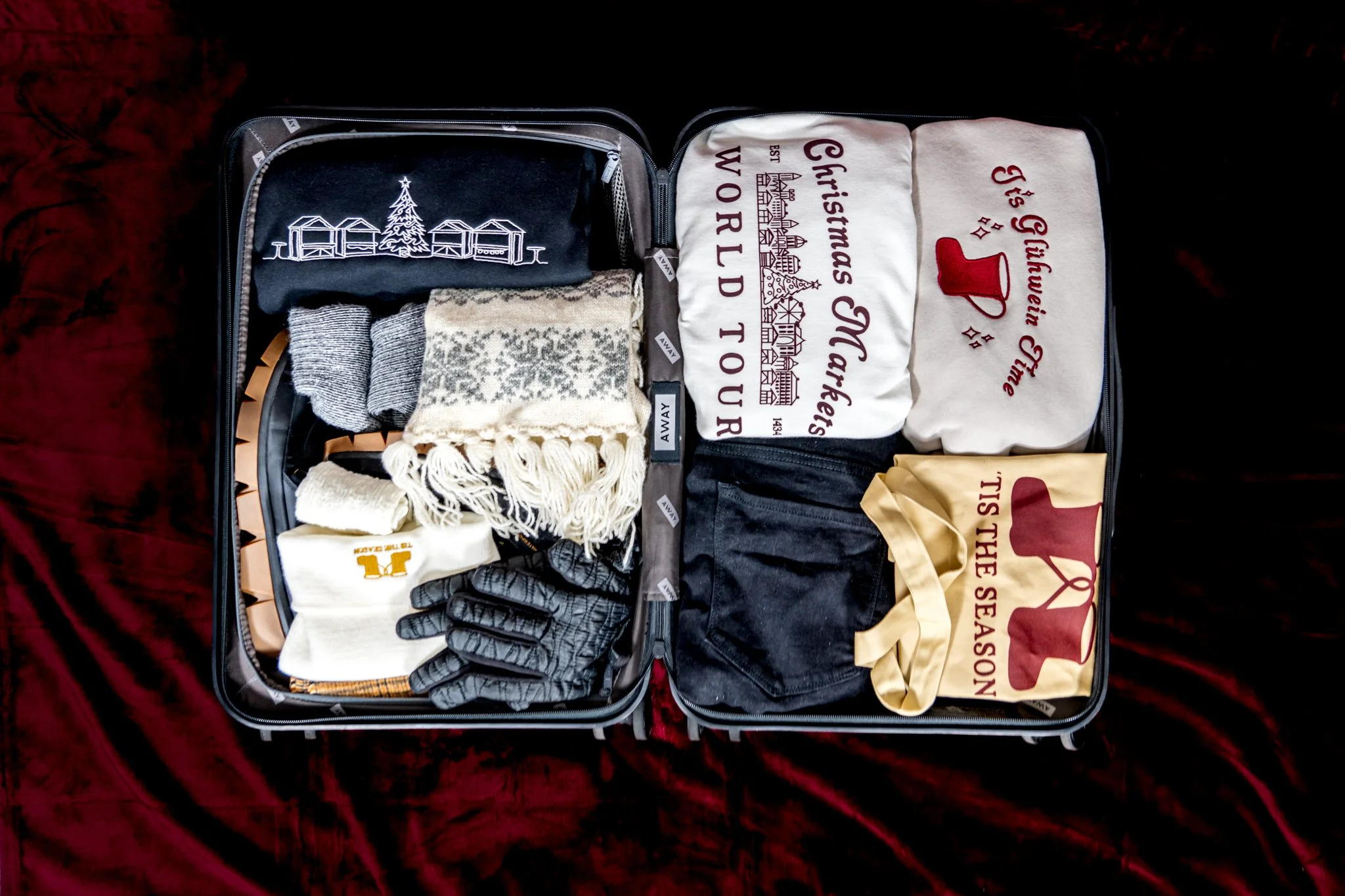 Open suitcase filled with winter clothing and accessories, including socks, gloves, scarves, and clothing with holiday-themed prints.