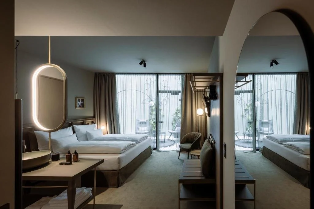 Hotel room with sliding glass door leading to a balcony, a bed with white linens, a vanity with a round mirror, a small sitting area with chairs, and a mirror reflecting the room.