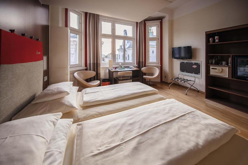 Hotel room with two beds, large windows with curtains, a small desk with a chair, a wall-mounted TV, and a shelving unit with appliances and decor.