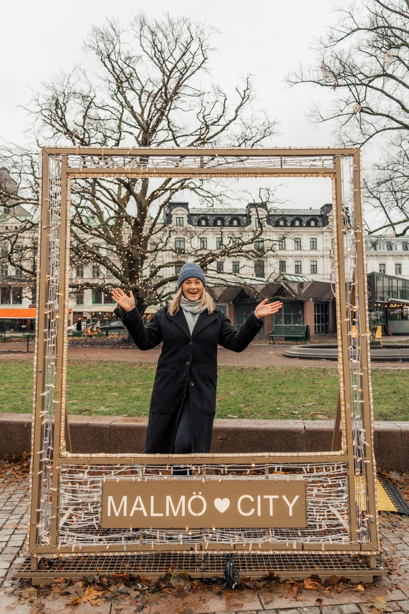 Woman in a black coat and gray beanie standing in front of decorative frame with Christmas lights that says 'Malmö City' in a park with trees and buildings in the background.