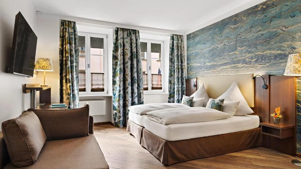 Comfortable hotel room with a large bed, decorative pillows, and a watercolor-style mural on the wall behind the bed. The room has large windows with patterned curtains, a wall-mounted TV, a small sofa, and bedside lamps.