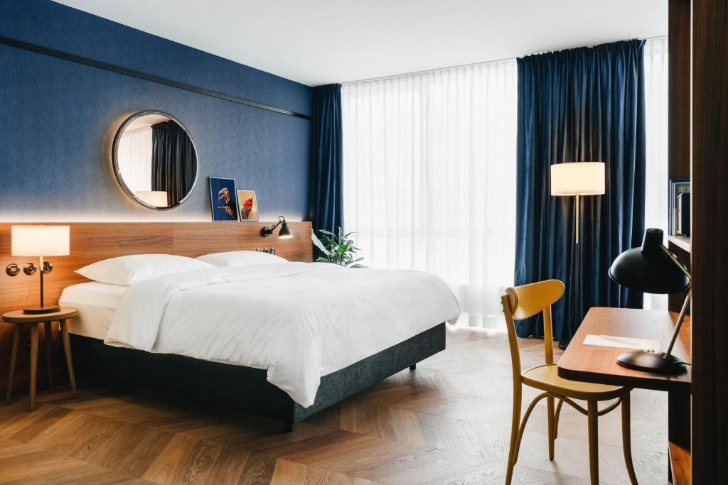 A modern hotel room with a large bed, wooden headboard, blue walls, and large window with white sheer and blue curtains. There is a nightstand with a lamp, some framed pictures, and a round mirror on the wall. A wooden desk with a yellow chair, a black desk lamp, and a book is also visible.