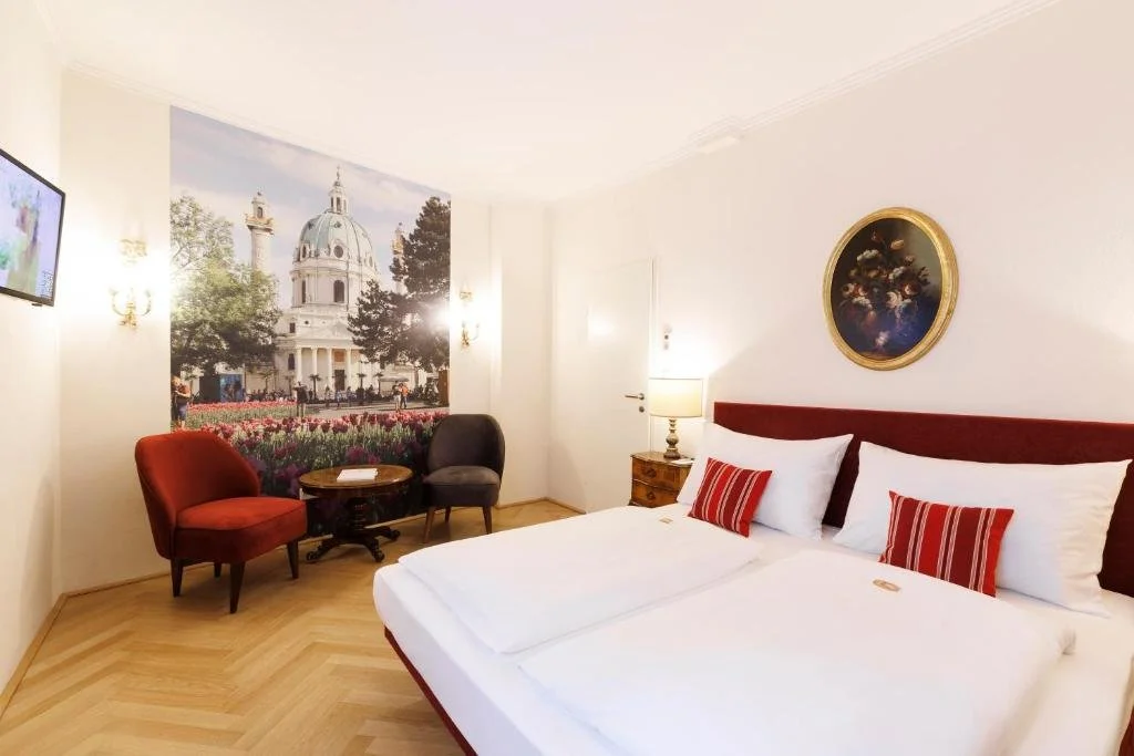 Hotel room with a large bed, red headboard, and three pillows with red and white stripes. A wooden nightstand with a lamp and a round painting above the bed. A corner with two chairs, a small table, and a large mural of a building with trees outside.