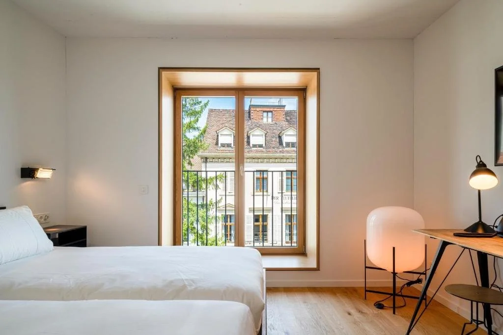 A minimalist hotel room with a large window, a bed, a small side table, a desk with a lamp, and a decorative floor lamp.
