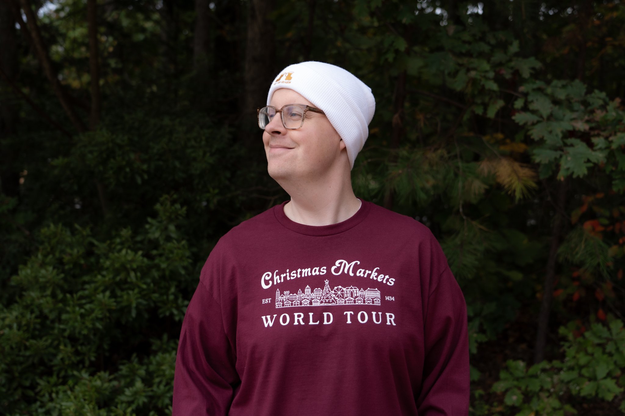 A person wearing a white beanie and glasses, smiling and looking to the side, standing outdoors with green trees in the background, dressed in a maroon long-sleeve shirt.