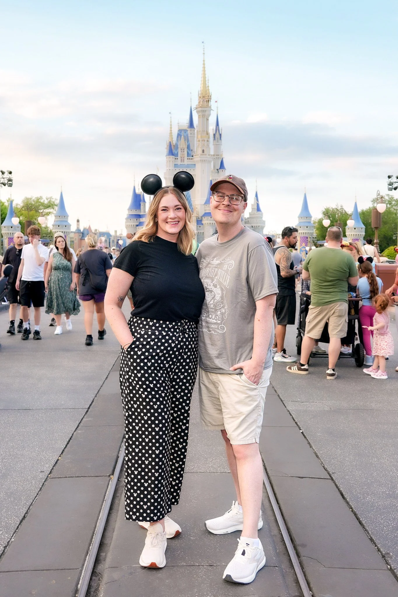 A smiling young woman wearing a black shirt, polka-dotted pants, white sneakers, and Mickey Mouse ears headband is standing next to a smiling young man wearing glasses, a gray T-shirt, beige shorts, and white sneakers. They are at a Disney theme park with Sleeping Beauty's Castle in the background and other visitors walking around.