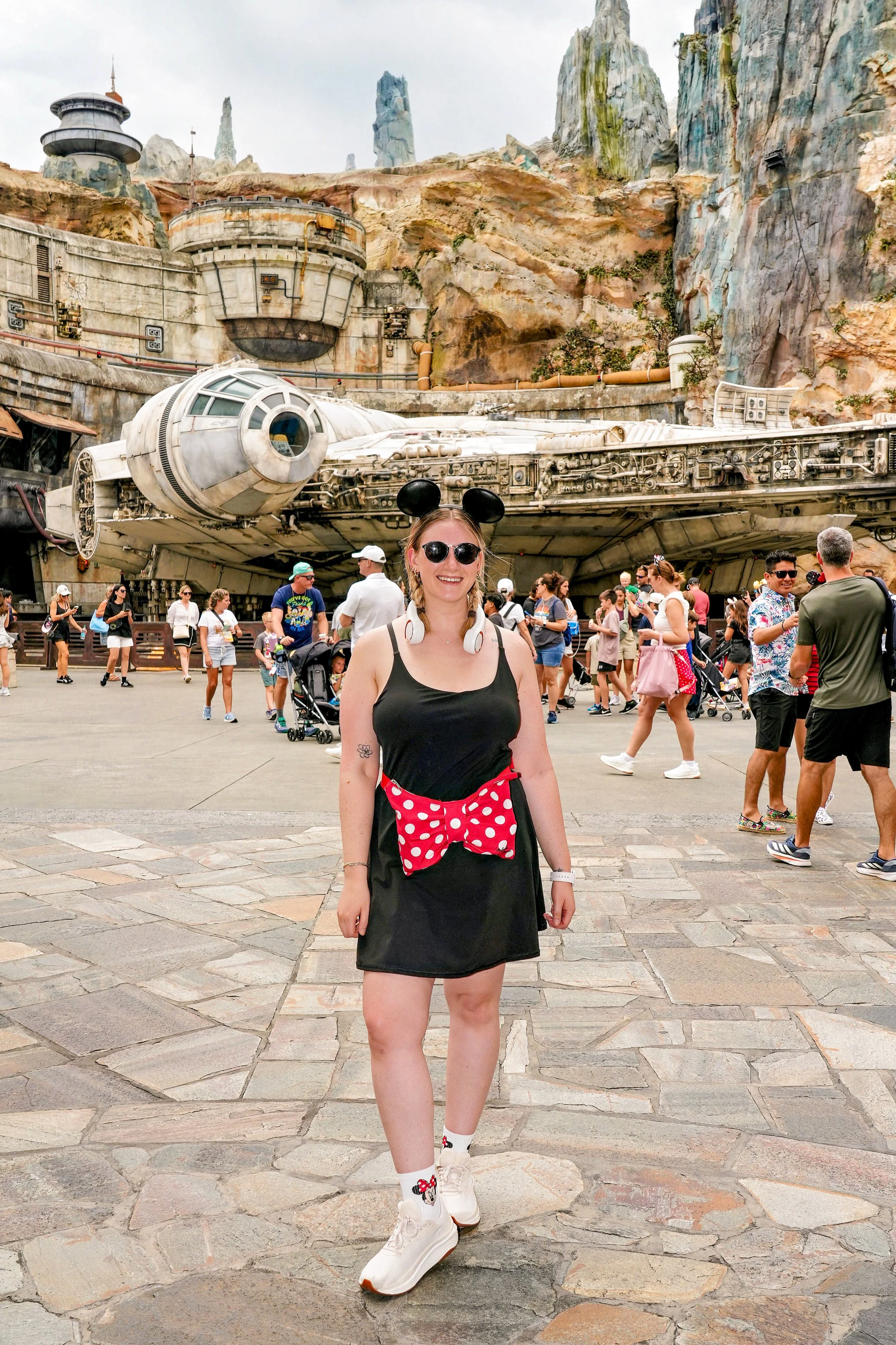 A young woman wearing black dress, Mickey Mouse ears, sunglasses, and white sneakers with Mickey Mouse socks, standing in front of the Millennium Falcon spaceship and rocky landscape at a Disney theme park.