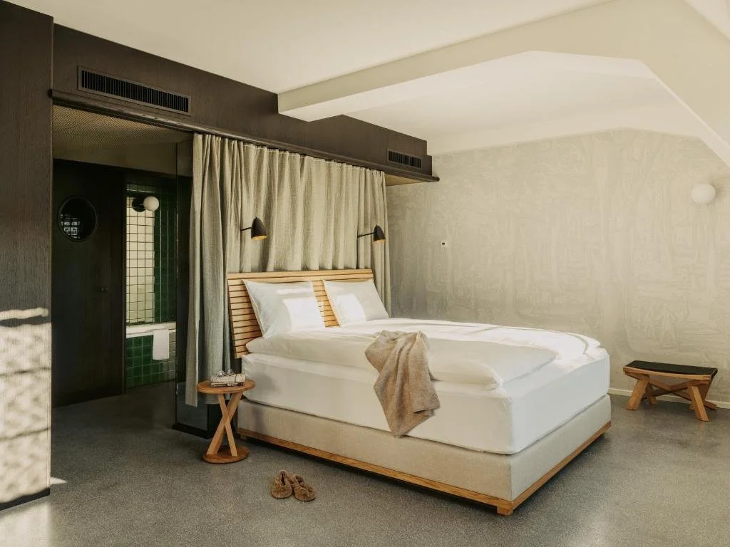 A modern hotel room with a large bed, beige linens, and a wooden headboard. There is a small side table with a towel and slippers, a bench at the foot of the bed, and a bathroom area with green tiles visible in the background.