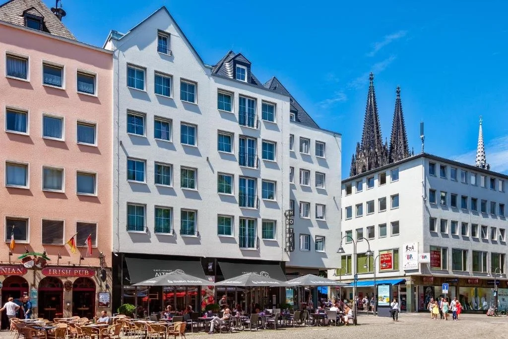 City street scene with multi-story buildings, a pub, hotel, outdoor dining area with tables and umbrellas, and people walking and sitting under a bright blue sky.