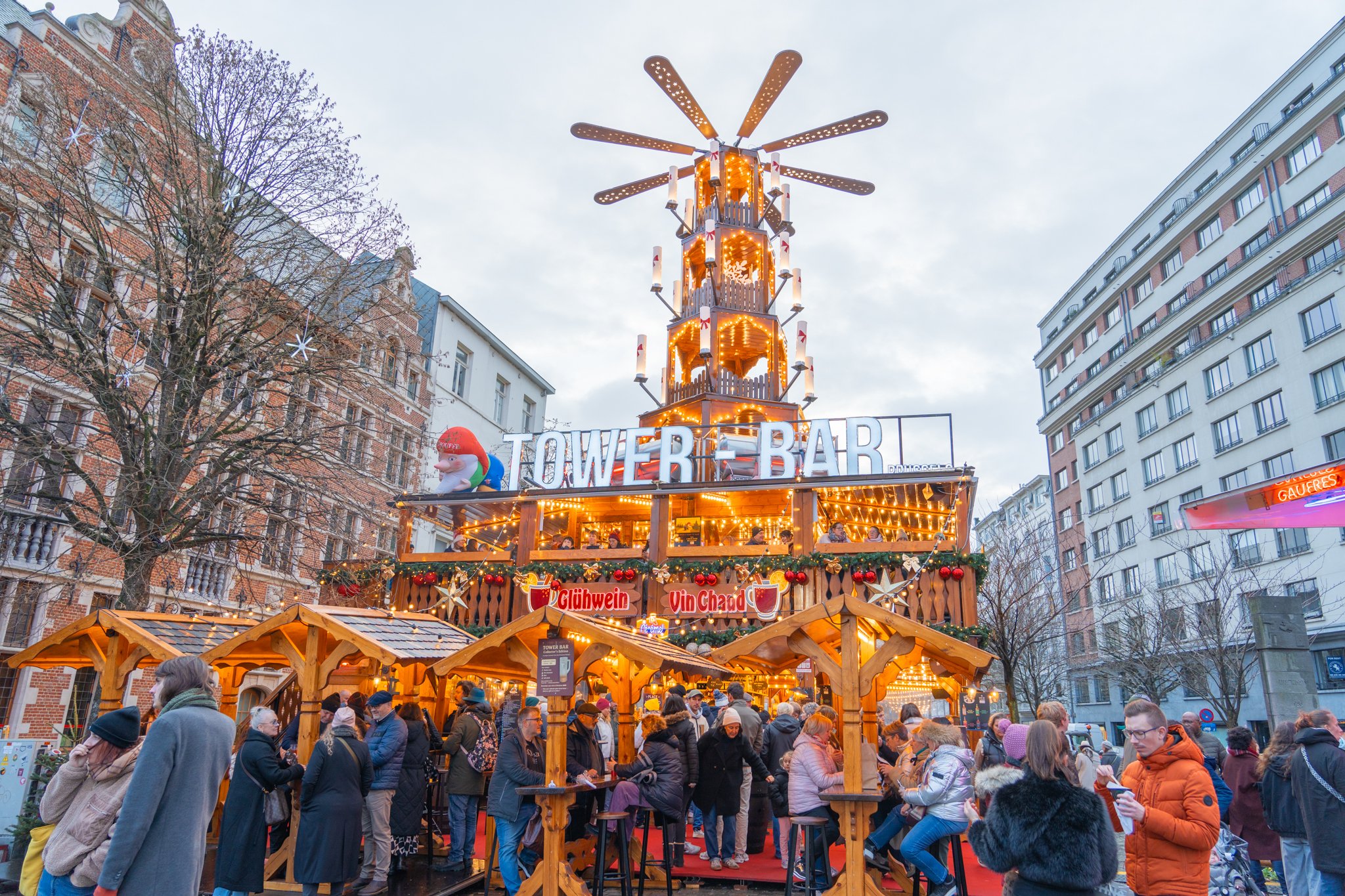 A bustling Christmas market scene featuring a large illuminated Tower Bar structure decorated with festive lights, garlands, and signs in German, with many people gathered around enjoying the holiday atmosphere in a city square surrounded by buildings.
