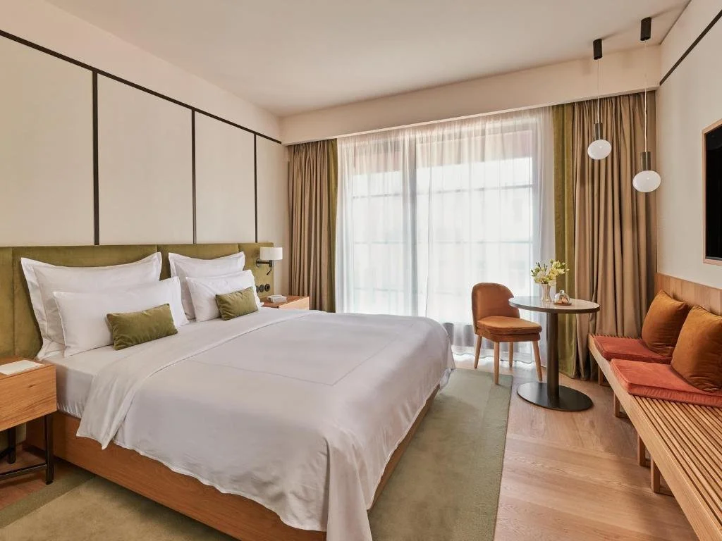 Modern hotel room with a large bed, green and white bedding, a small round table with a flower vase, a brown chair, a wooden bench with cushions, and a large window with curtains.