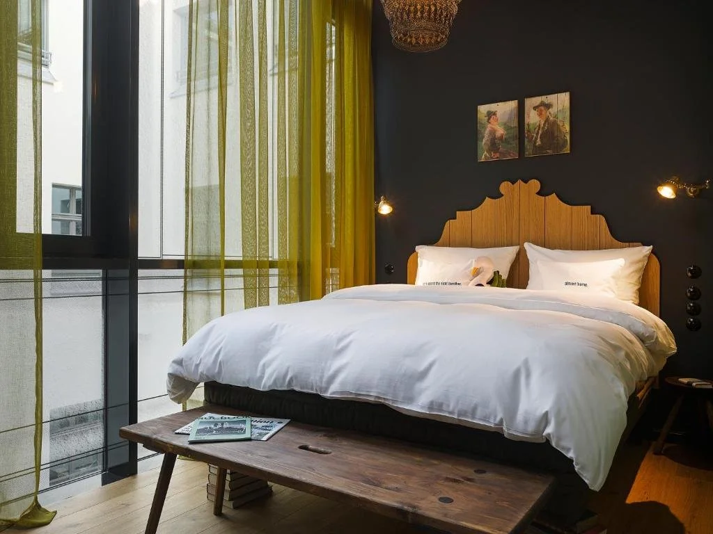 A modern bedroom with a large bed covered in white linens and two pillows. The bed has a wooden headboard, flanked by two wall-mounted lights. Above the bed are two small paintings. To the left, large windows with sheer yellow curtains let in natural light, and there is a wooden bench at the foot of the bed with magazines on it.