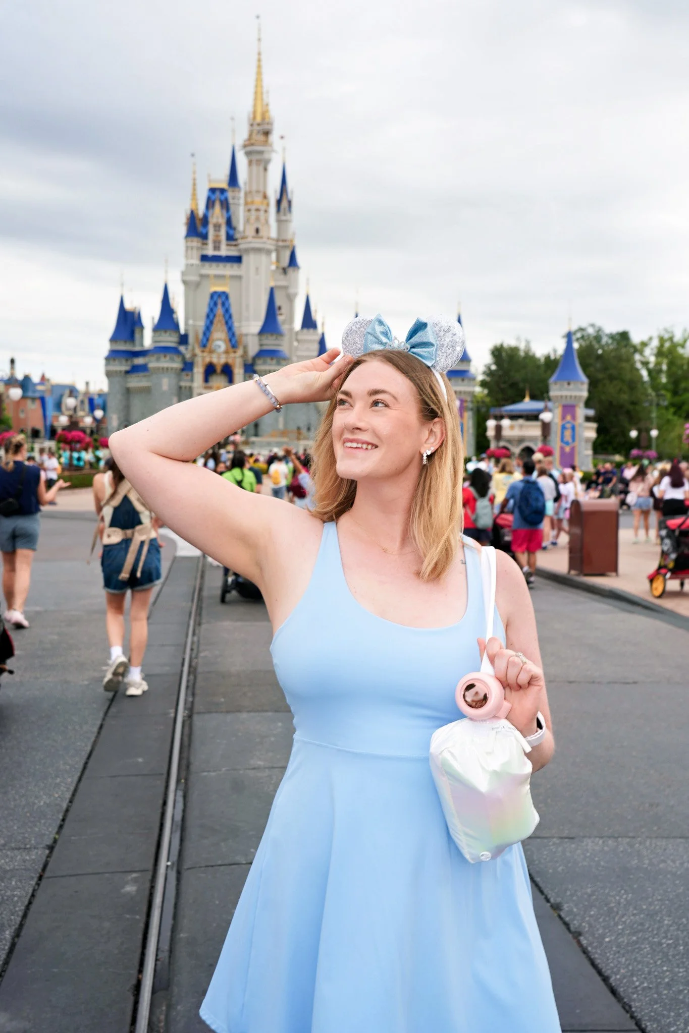 A young woman wearing a light blue dress and silver Minnie Mouse ears headband standing in front of Sleeping Beauty Castle at Disneyland, smiling and holding her belongings.