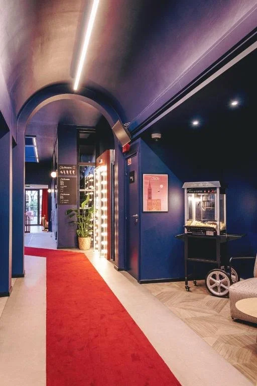 Indoor hallway with dark blue walls, red carpet, potted plant, illuminated entrance, and a popcorn machine on a cart.