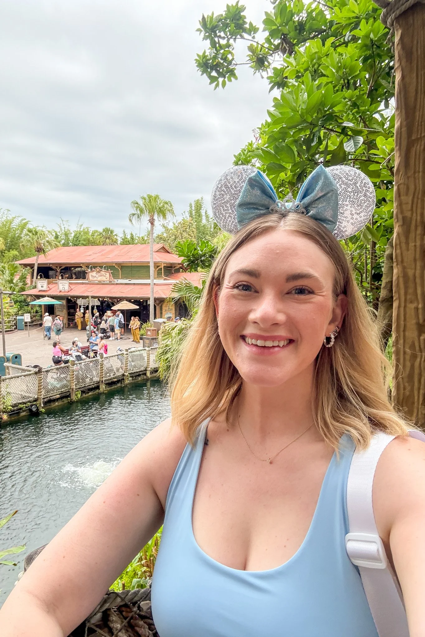 A woman wearing a blue tank top and a Disney Minnie Mouse ears headband taking a selfie outdoors near a water feature with a crowded park in the background, including a building with a red roof and palm trees.