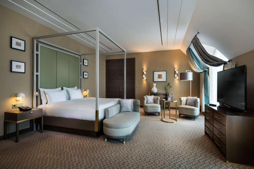 A modern hotel room featuring a large four-poster bed with white linens, surrounded by wall art and lamps. The room has beige walls, a carpeted floor, a sitting area with two chairs and a small table, and a large flat-screen TV on a wooden dresser near a window with draped curtains.