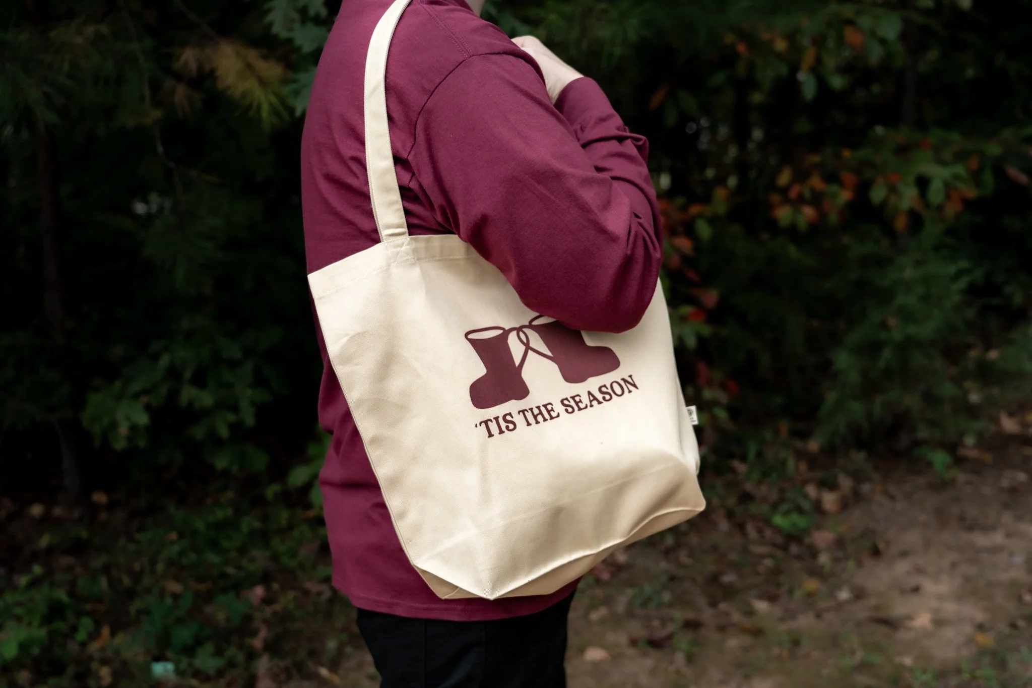 Person wearing a maroon long sleeve shirt carrying a beige tote bag with burgundy text and graphic, standing outdoors near bushes.