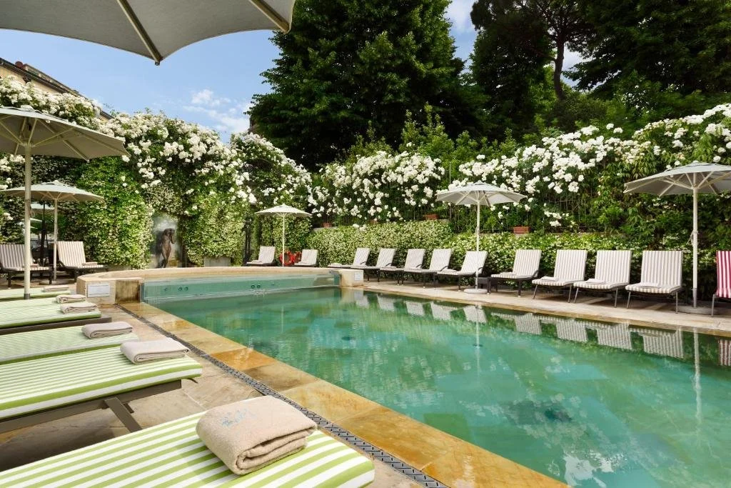 A swimming pool surrounded by lounge chairs and umbrellas, with flowering bushes and trees in the background.