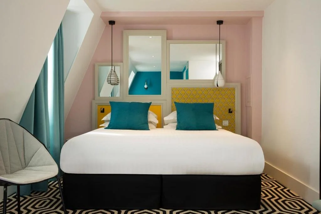 Modern hotel room with a bed, teal pillows, yellow headboard, blue accent wall, and patterned black and white floor.