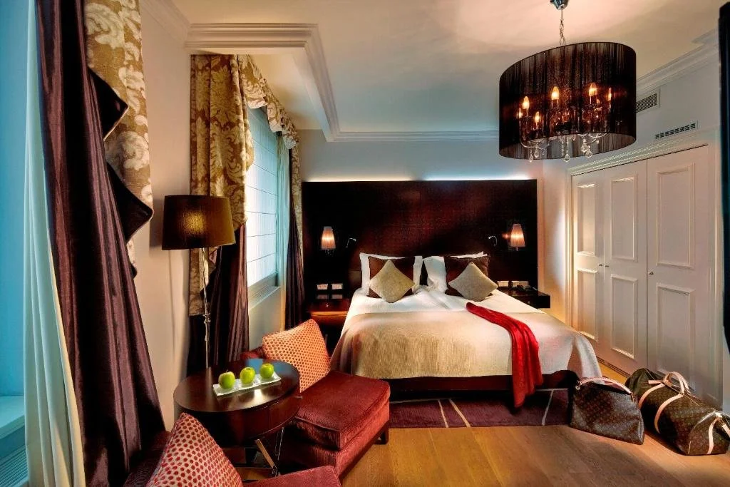 A luxurious hotel bedroom with a large bed, dark headboard, and pillows. There are two bedside lamps, a window with patterned curtains, a small seating area with a wooden table, armchair, and green apples. A chandelier hangs from the ceiling, and there are duffel bags on the floor near a white closet.