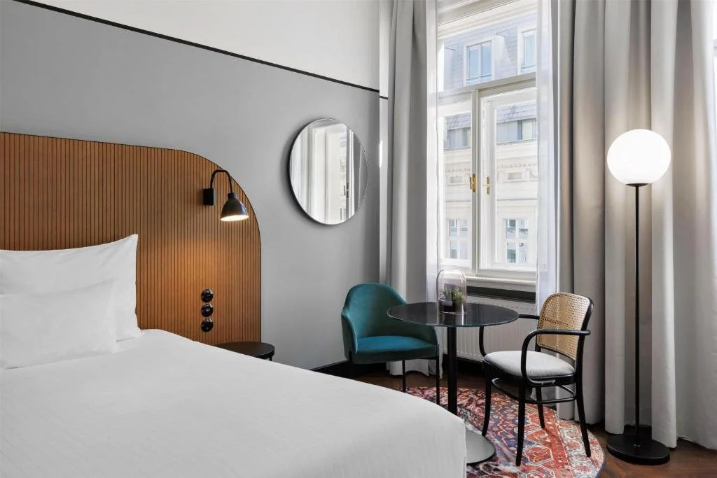 Modern hotel room with a bed, wood headboard, black reading lamp, round mirror on a white and gray wall, a small sitting area with a teal armchair, a glass terrarium with plants, a black dining table, and two chairs, near a large window with white curtains and a tall floor lamp.