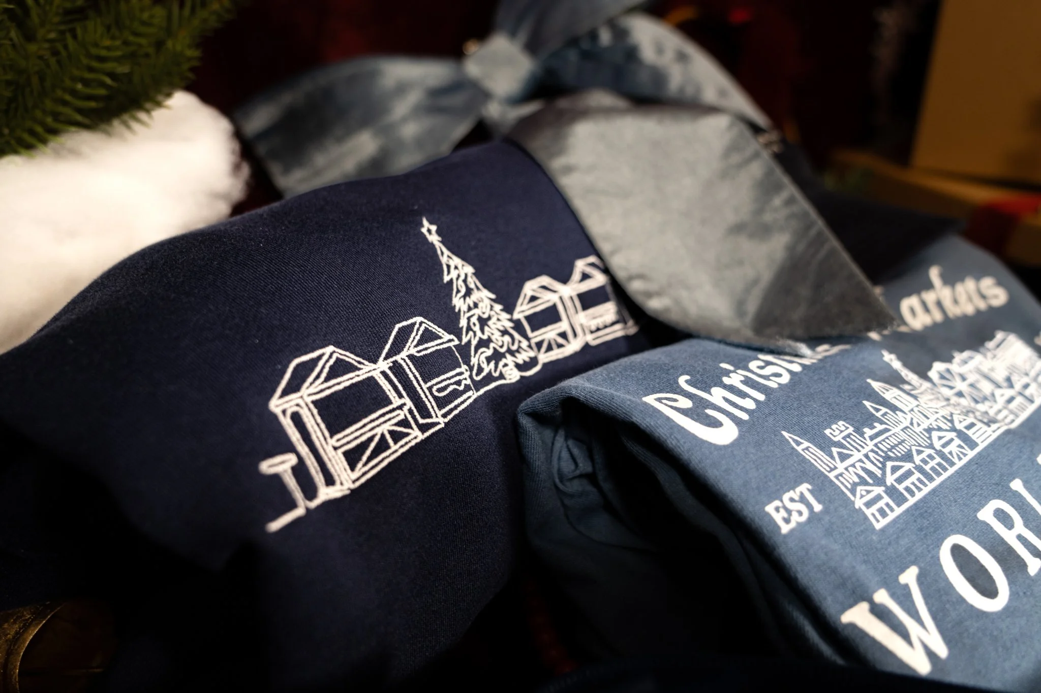 Folded Christmas-themed clothing items featuring white line art of houses and trees on dark fabric, with text indicating Christmas village and establishments.