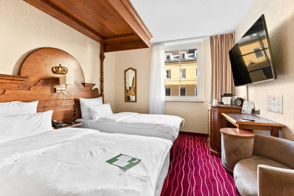 Hotel room with two beds, wooden headboard with a crown decoration, a window with a view of a yellow building, a flat-screen TV mounted on the wall, a small desk with a chair, and a chair with a blanket, with pink carpet and neutral-colored walls.