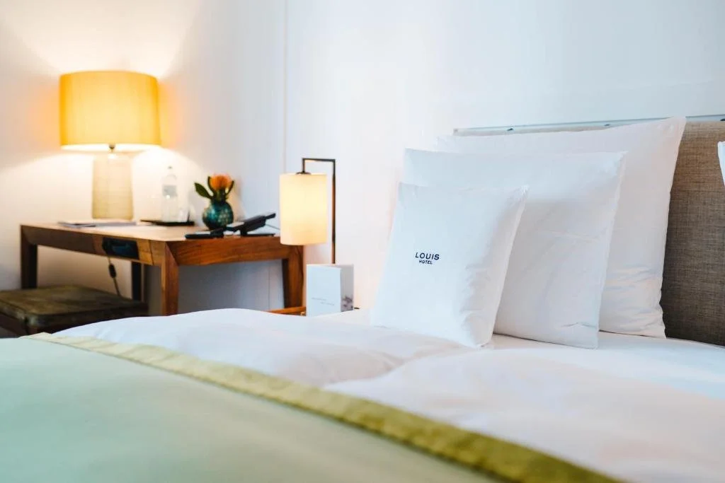 A neatly made hotel bed with white pillows and sheets, a bedside lamp, and a wooden desk with a vase and a phone, in a well-lit hotel room.