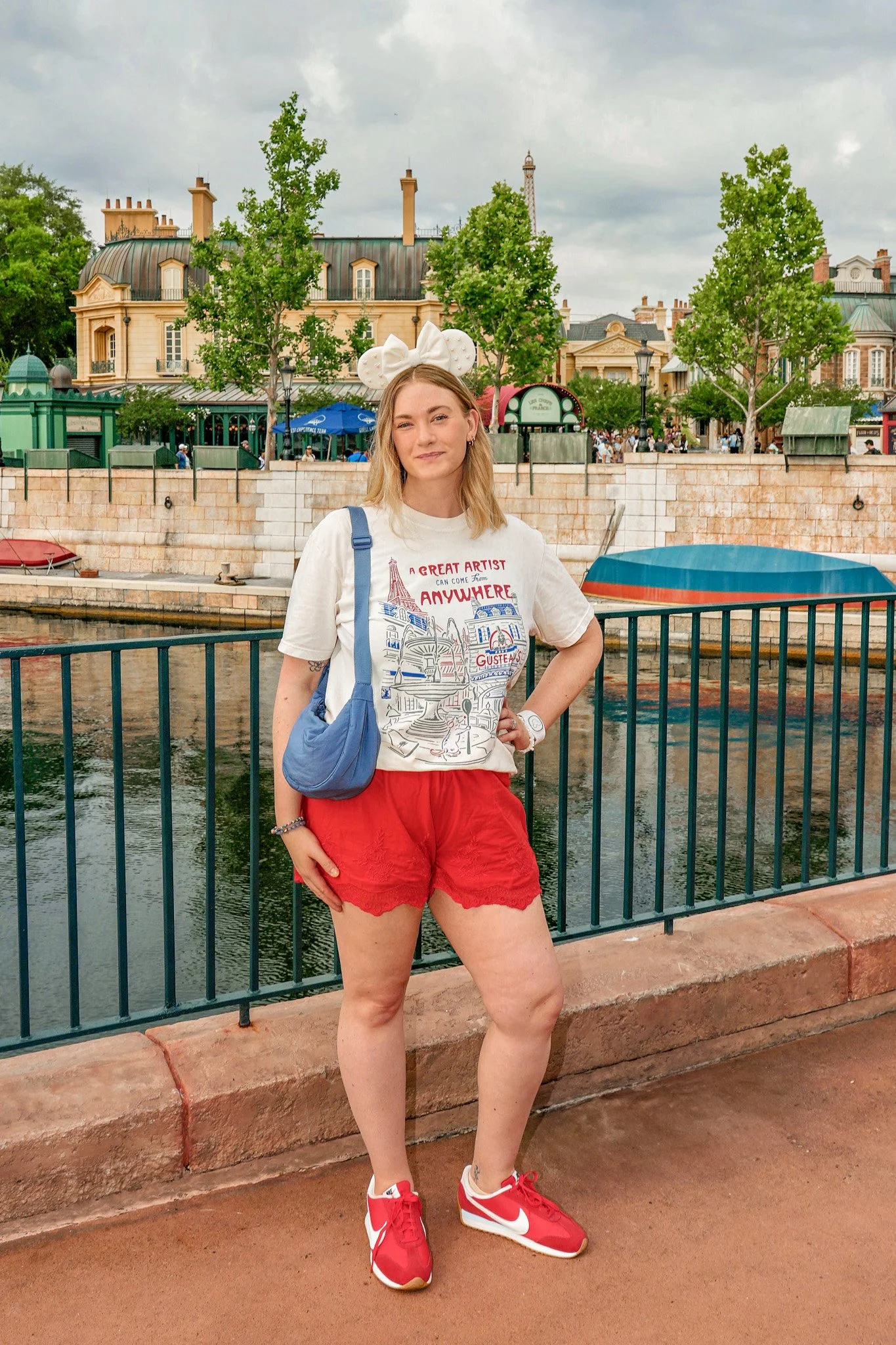 A young woman standing by a canal with green trees and historic buildings in the background. She is wearing a white T-shirt with red and blue graphics, red shorts, red and white sneakers, and a headband with white, circular ears.