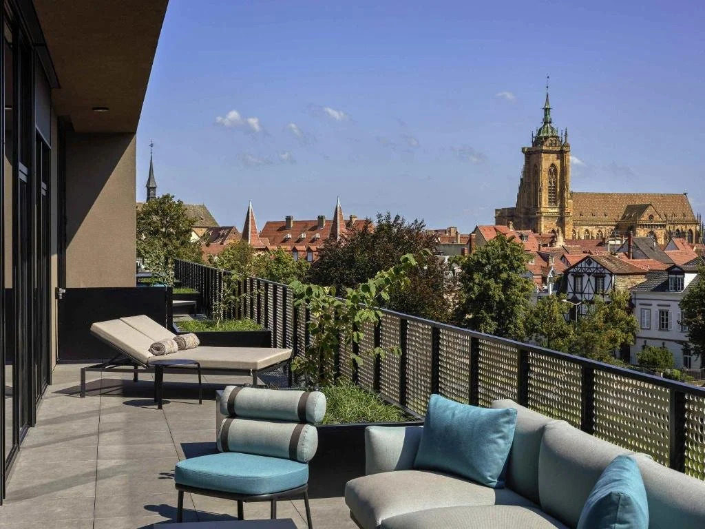 Balcony with lounge chairs and a sofa overlooking a cityscape with historic buildings and church towers.