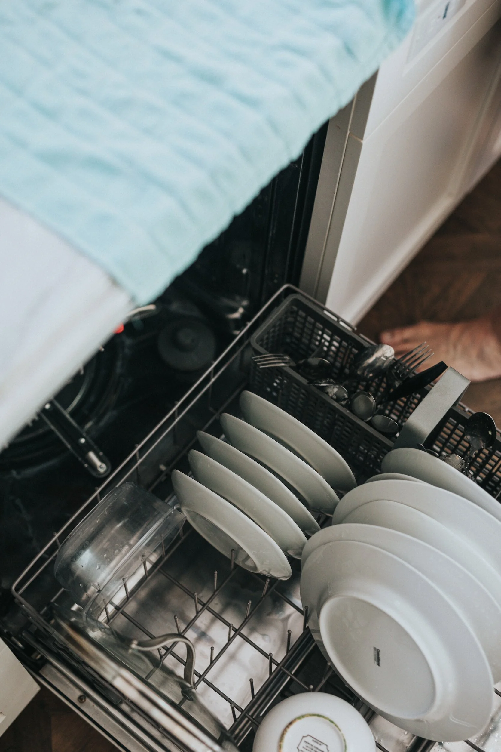 Open dishwasher with clean white dishes and utensils inside, covered with a blue cloth.