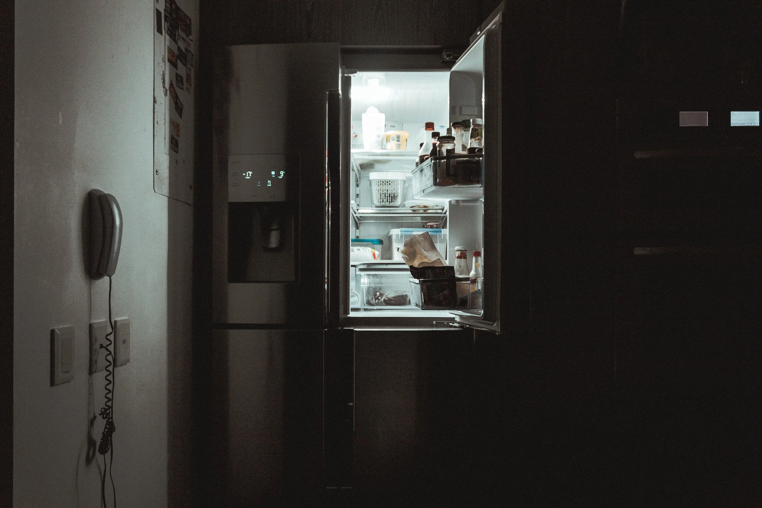 Open refrigerator in dimly lit kitchen, showing interior shelves stocked with various bottles and food containers; a wall phone is visible nearby.
