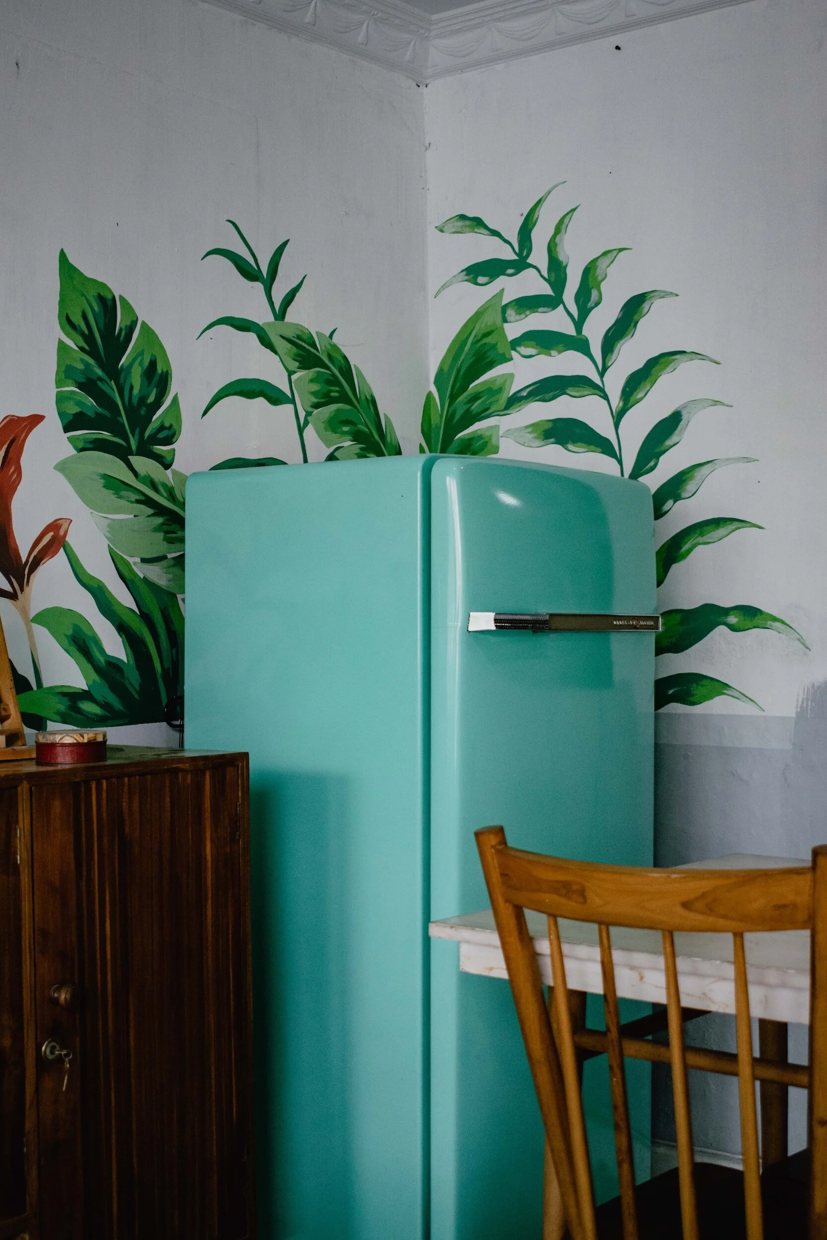 A retro blue fridge against a wall with leaf-themed wallpaper, next to a wooden cabinet and chair.