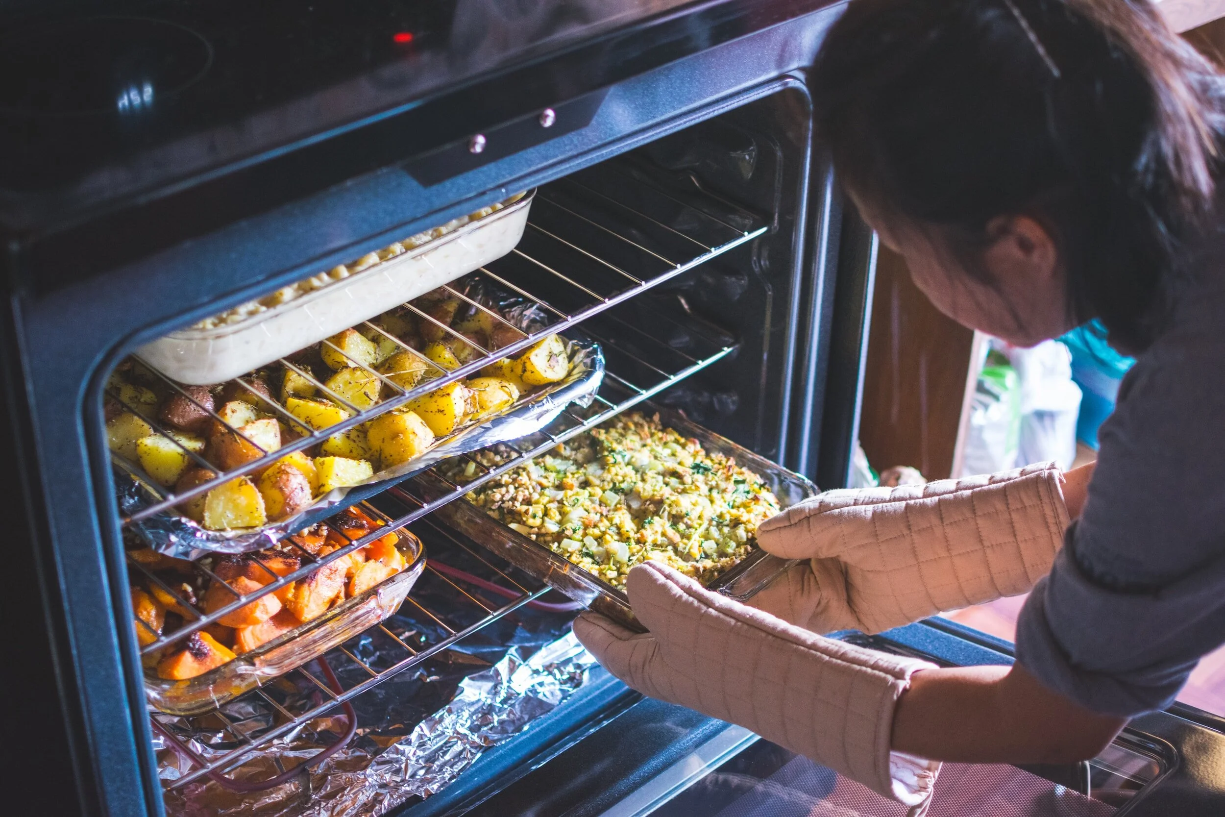 Person placing a casserole dish into an oven with other dishes, including roasted potatoes and vegetables.