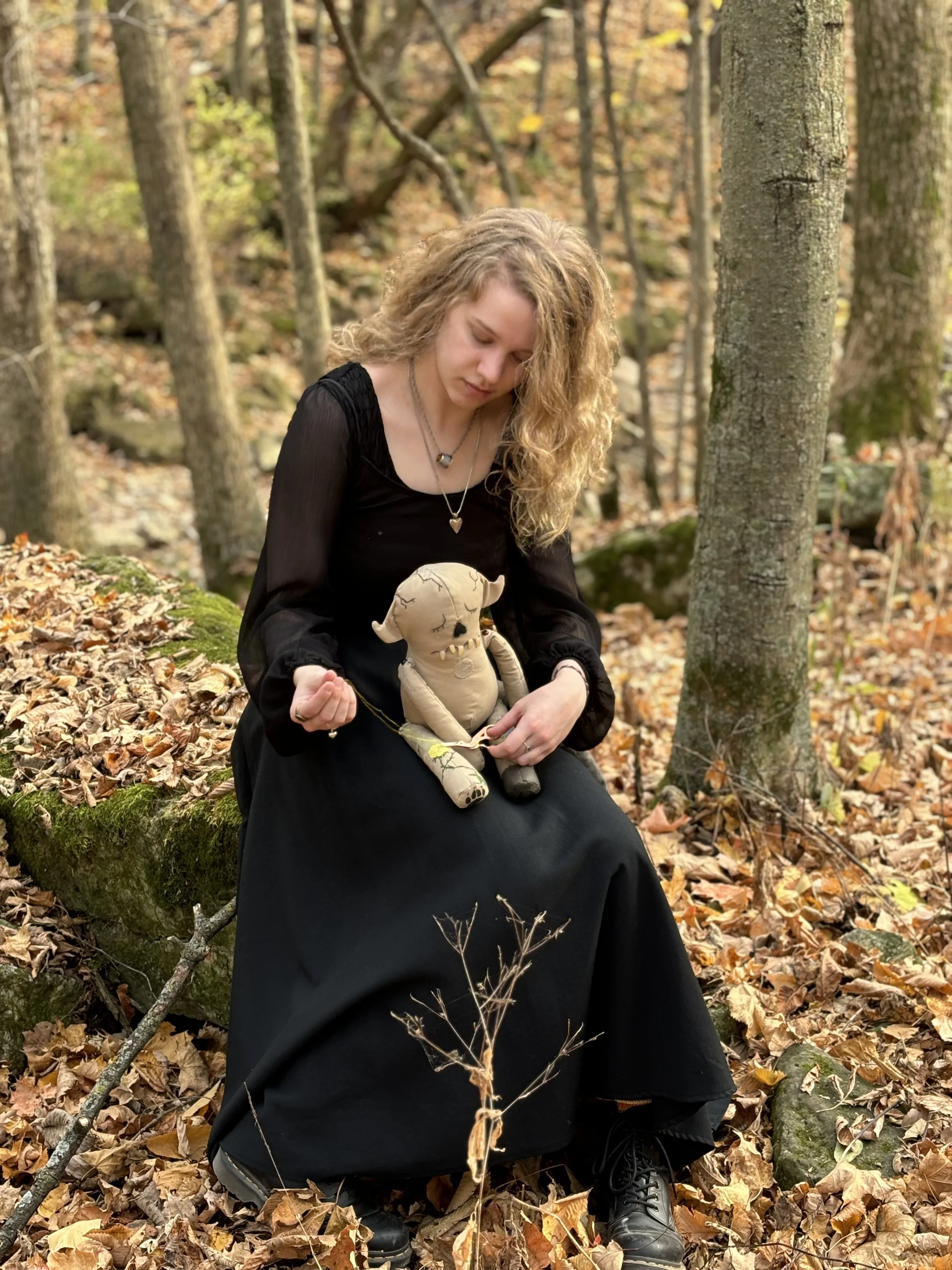 A young woman in a black dress sits in the woods sewing a small creature in her lap.