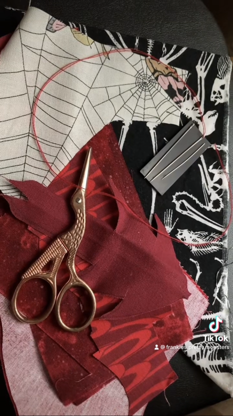 A pair of rose gold sewing scissors rests on a pile of red, black, and cream patterned fabric next to three silver sewing needles.