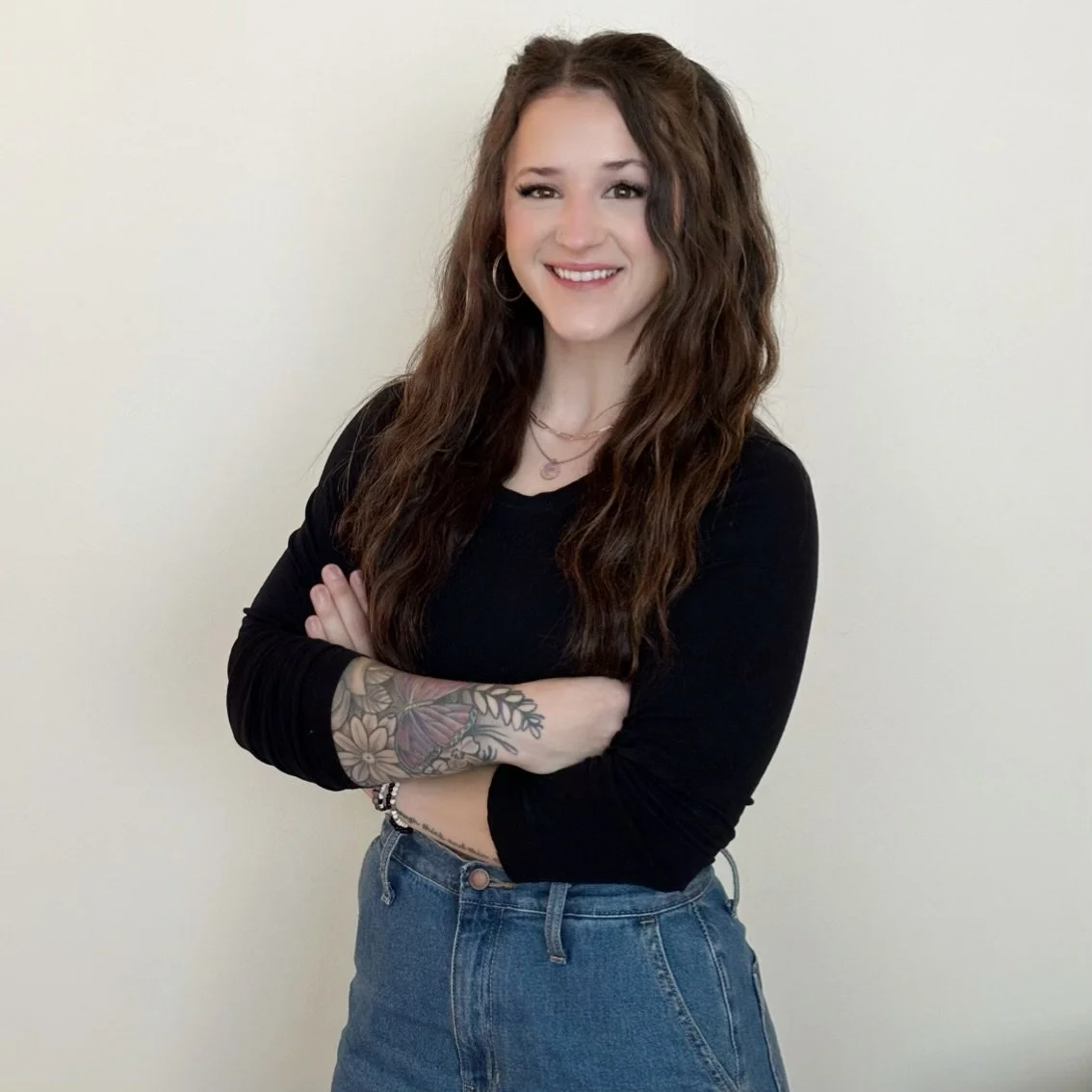 A young woman with long, wavy brown hair, wearing a black long-sleeve shirt and blue jeans, standing against a plain light-colored wall, smiling with arms crossed and displaying a floral tattoo on her left forearm.