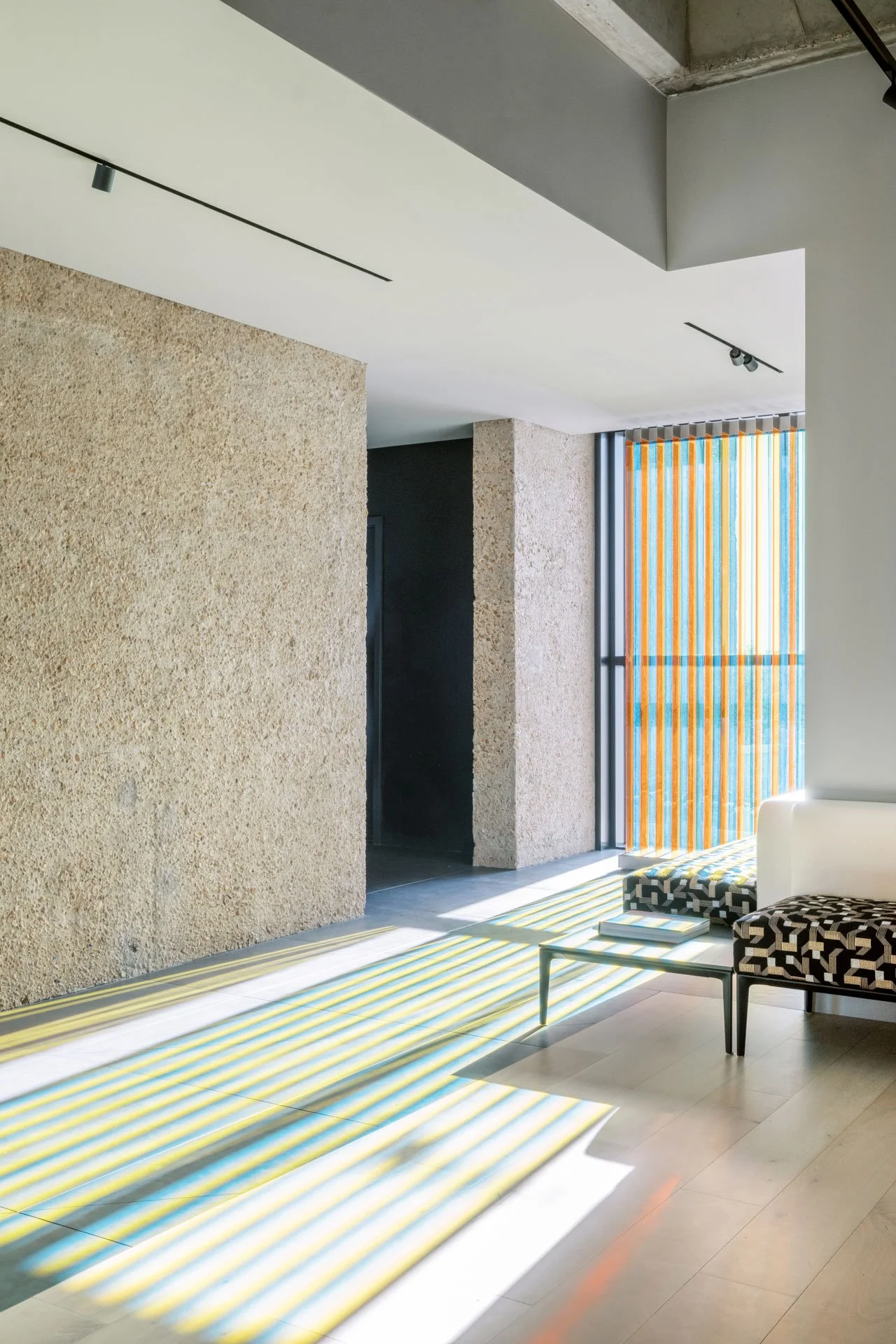 Modern interior with textured beige walls, a black door, a white ceiling with track lighting, colorful vertical blinds casting striped shadows on the floor, a white sofa, and patterned ottomans.