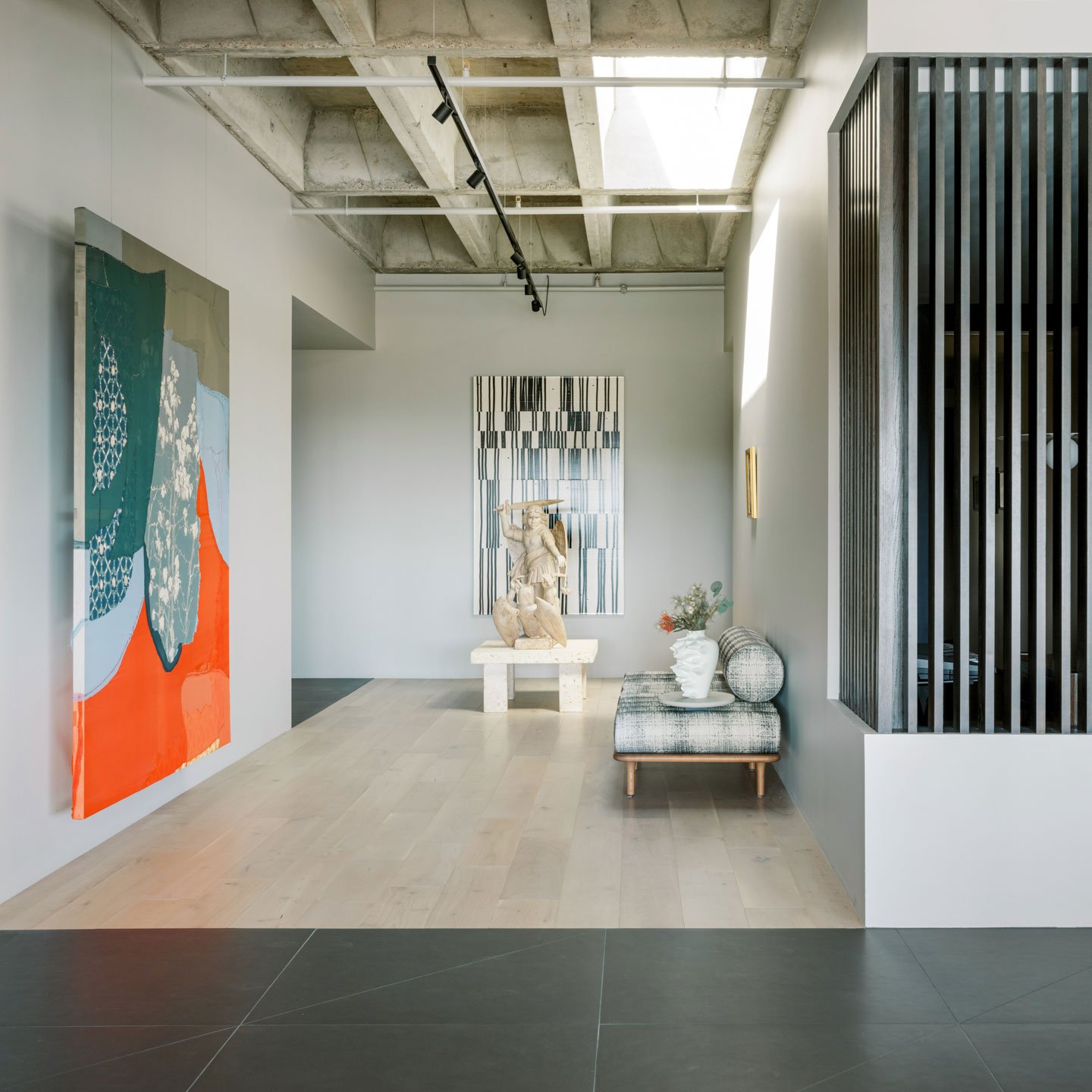 Modern art gallery interior featuring large paintings on the walls, a sculpture on a pedestal, a bench with decorative vases, and a concrete ceiling with track lighting