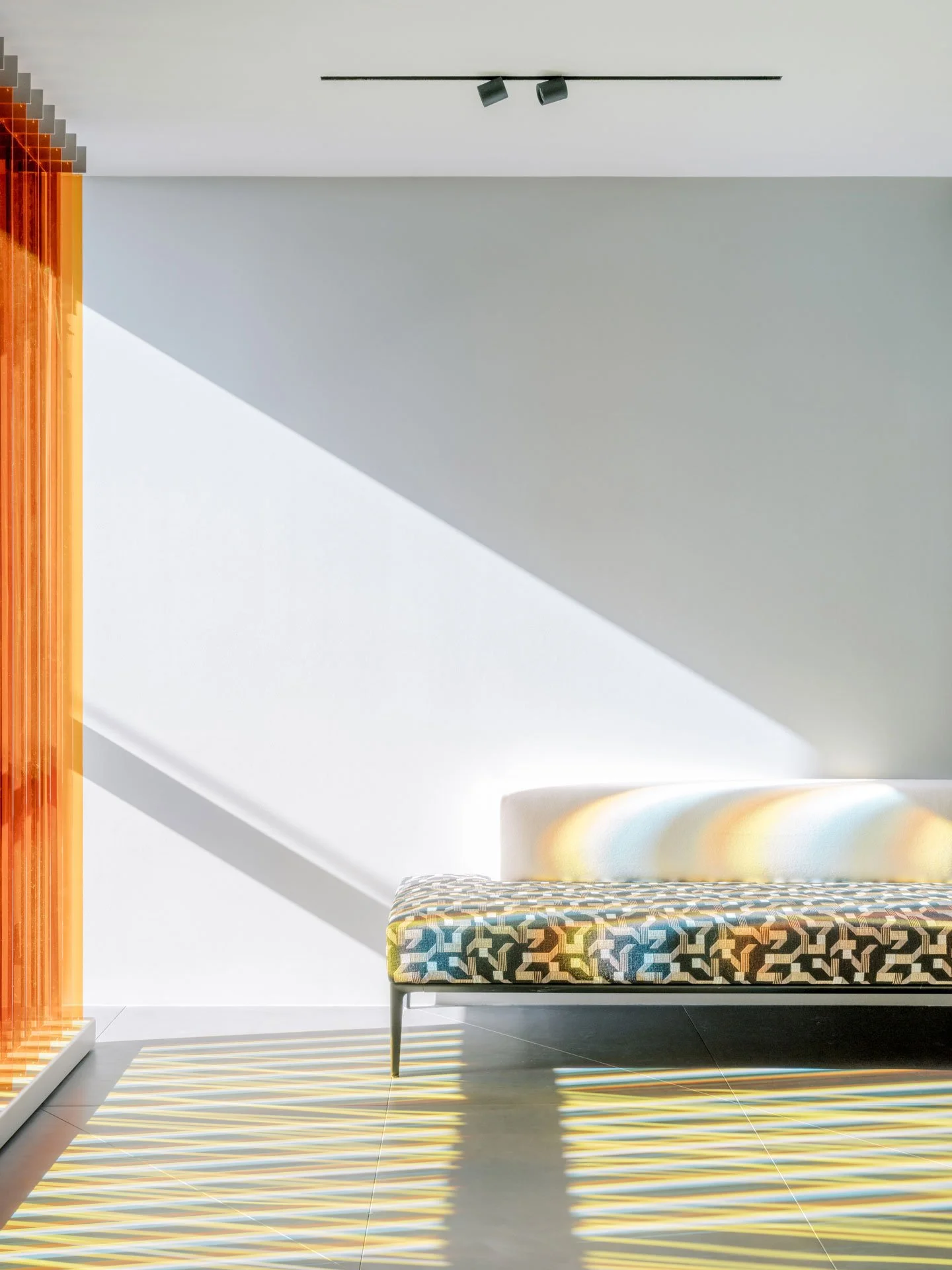 Modern living room interior with a patterned sofa, sunlight creating striped shadows on the tiled floor, and a white wall with a beam of natural light casting a shadow.