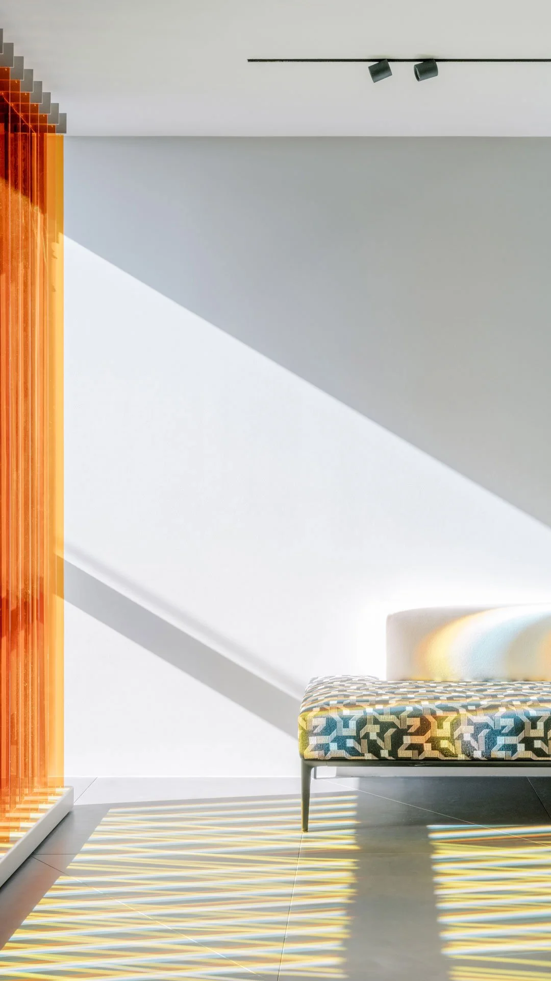 Modern interior with geometric patterned sofa, orange wooden vertical slats, and sunlight creating striped shadows on tiled floor.