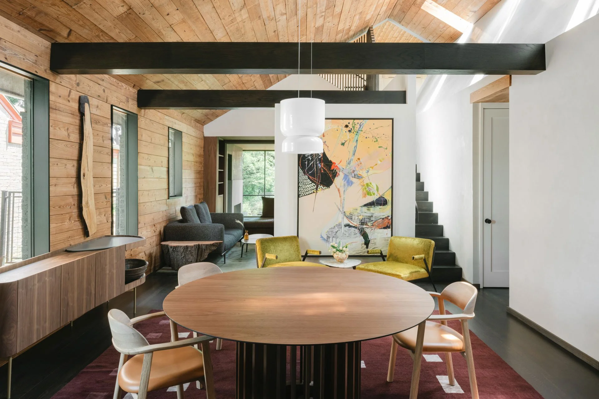Interior of a modern dining and living room with wooden walls and ceiling, featuring a round dining table with six chairs, a black sofa, abstract artwork on the wall, and a staircase leading to an upper level.
