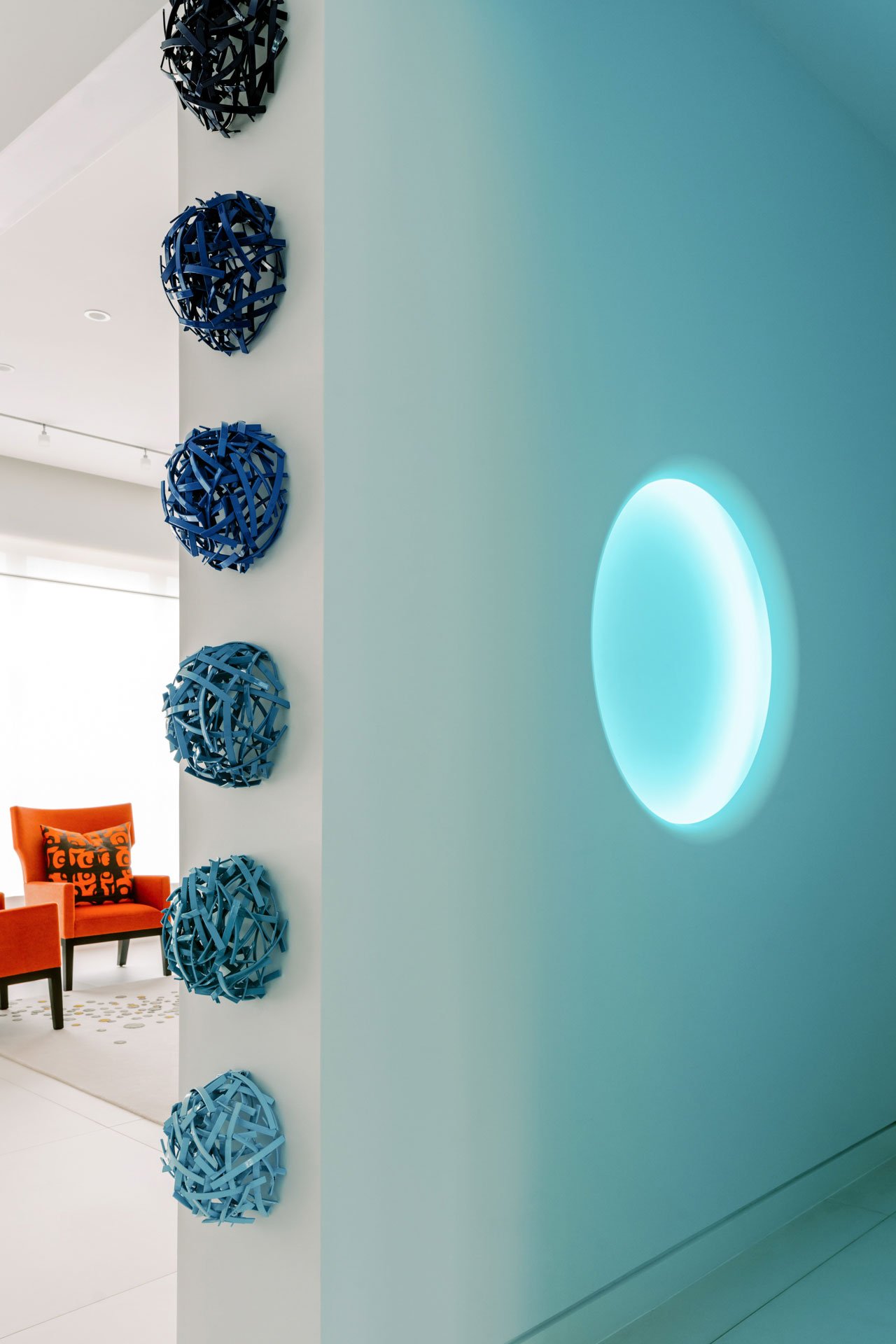 A modern interior with a white wall decorated with six blue spherical sculptures arranged vertically, and a glowing circular light fixture on the right. In the background, there are orange armchairs with a patterned pillow and a light-colored rug.