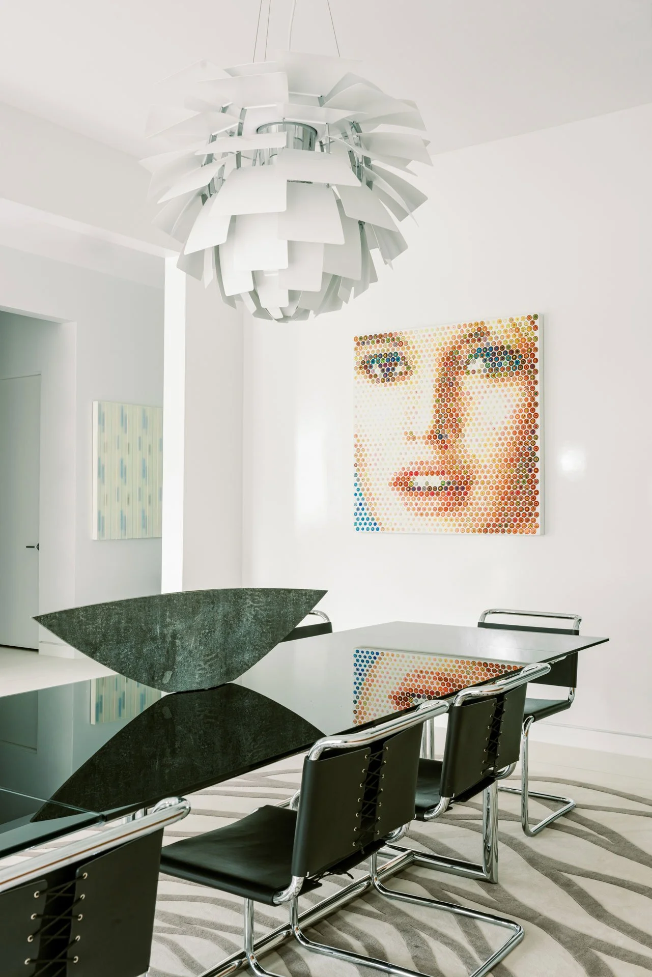 Modern dining room with black glass table, four black chairs with chrome frames, abstract white pendant light, colorful portrait pixel art on white wall, and a striped rug.
