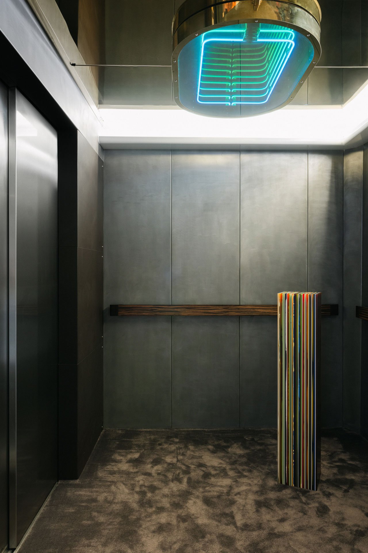 Empty elevator with dark walls and a colorful striped sculpture on the floor, illuminated by a neon blue and green lighting fixture on the ceiling.