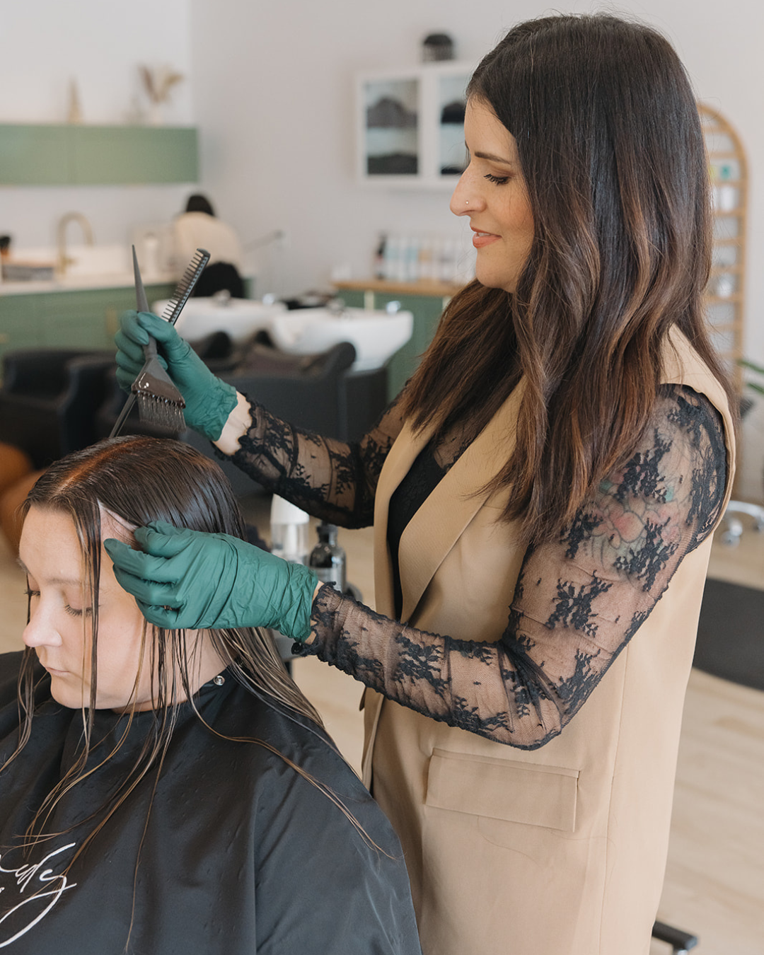 A hairstylist with long dark hair, wearing a beige vest and teal gloves, styling a woman's hair in a salon.