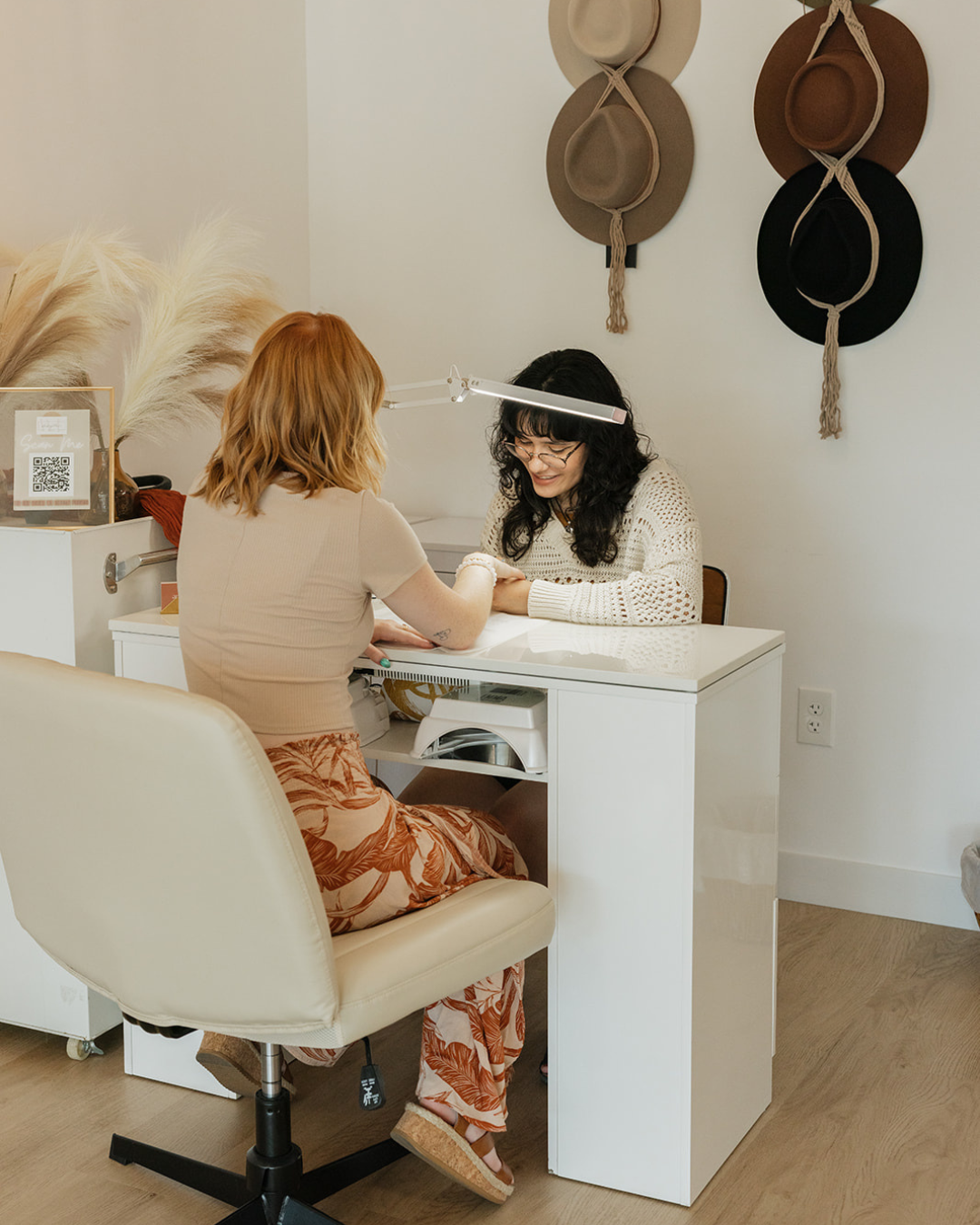 A woman with red hair getting a manicure from a woman with black hair and glasses at a white nail salon desk. The woman receiving the manicure is smiling, and a bright LED light is illuminating the scene. Behind them are beige, brown, and black hats hanging on the wall.